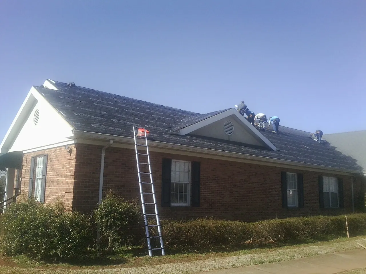 Crew tearing shingles off a brick ranch roof with a ladder set — Emergency Roof Repair work by our crew in Gum Spring